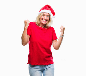 中年金发女人戴着圣诞帽，在孤立的背景下，非常高兴和兴奋地做着胜利者的手势，手臂抬起，微笑着，尖叫着，争取成功。 庆祝概念。