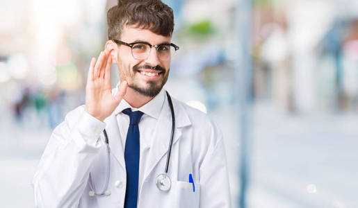 年轻的医生穿着医院外套，在孤立的背景上微笑，用手捂住耳朵听谣言或流言蜚语。 耳聋概念。