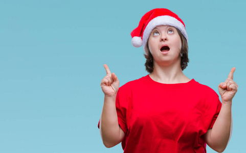 年轻的成年妇女戴着圣诞帽，戴着孤立的背景，惊讶地抬头，用手指和举起的手臂指着。