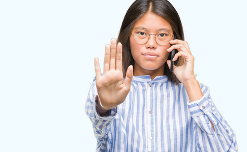 年轻的亚洲女人在电话中，在孤立的背景下，张开手做停止标志，严肃而自信的表情防御手势