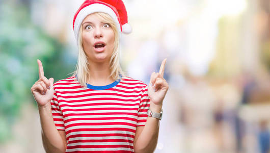 年轻漂亮的金发女人戴着圣诞帽，戴着孤立的背景，惊讶地抬头，用手指和举起的手臂指着。