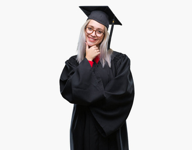 年轻的金发女人穿着研究生制服，在孤立的背景下，自信地看着相机，微笑着交叉的手臂和举起的手在下巴上。 积极思考。