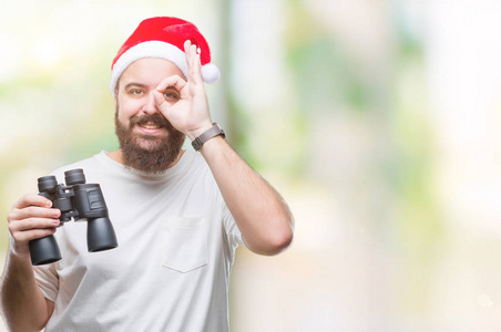 年轻的白种人，戴着圣诞帽，戴着双筒望远镜，在孤立的背景上看着，快乐的脸，微笑着，做着确定的标志，用手看着手指