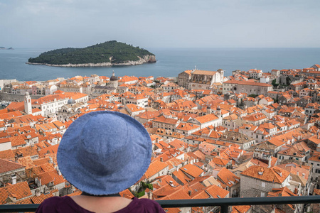 白种人女游客戴着蓝色帽子站在杜布罗夫尼克的旧城墙上，欣赏着克罗地亚下面历史名城的景色