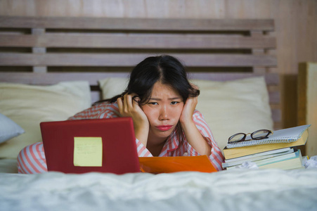 年轻的沮丧和疲惫的亚裔美国大学生女孩感到不知所措，紧张地准备考试，用笔记本电脑学习，在家里的床上过度劳累