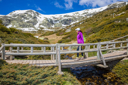 穿着紫色夹克衫的女子登山者穿过一座木桥，越过一条小溪，看着皮卡拉山地平线上的雪山西班牙马德里公园