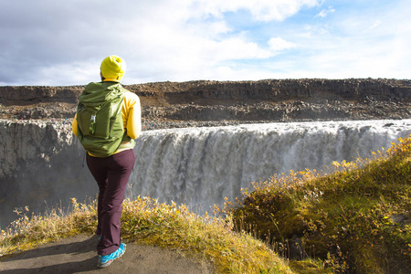 一位穿着黄色衣服的年轻女子站在悬崖边，俯瞰着冰岛巨大的Detifoss瀑布。 晴朗的暑假