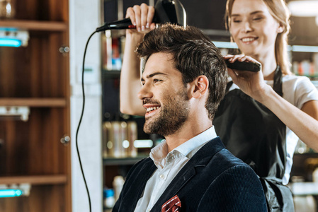 微笑的年轻发型师在美容院给英俊的快乐男人烘干头发