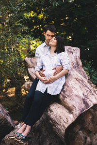 美丽的怀孕夫妇在森林里放松，坐在树茎上拥抱和亲吻，背景是绿叶。