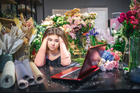担心的年轻花商牵着手在头上看着笔记本电脑。桌子上有纸卷。她身后有许多五颜六色的花朵和植物