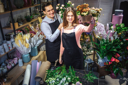 年轻的女花和雄性花店站在满屋子的花和植物里。她抱着磨练。他们拿了自拍。人们微笑着摆姿势