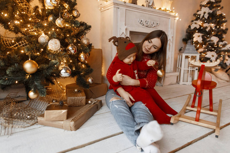 欢快可爱的年轻女人妈妈穿着一件红色毛衣，坐在工作室的地板上，在玩具和礼物的背景下，和一个穿着时尚西装的可爱的婴儿玩耍。 圣诞气氛
