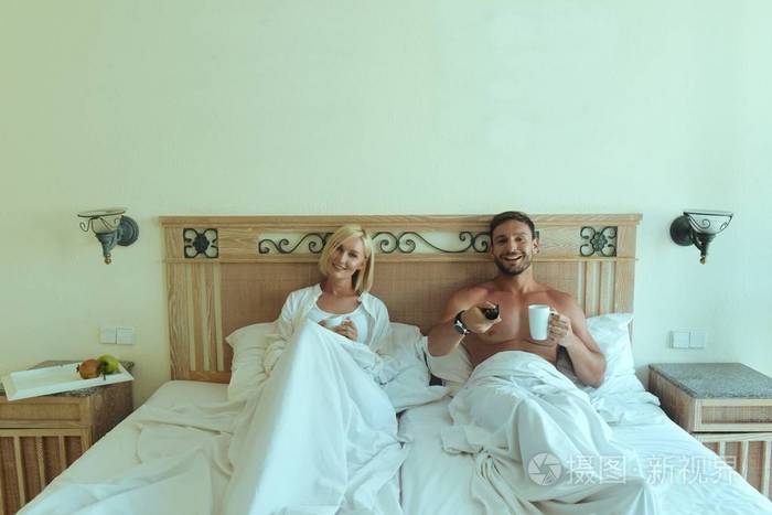 帅哥和美女躺在床上玩,在卧室里看电视,迷人的夫妻在性生活后放松