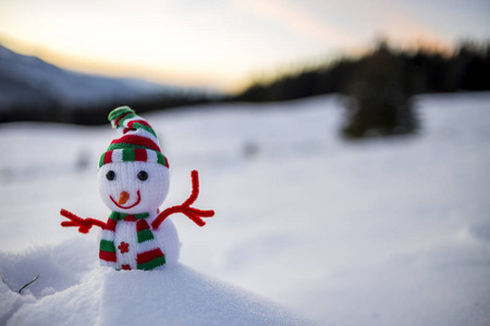 小有趣的玩具婴儿雪人在针织帽子和围巾在大雪户外模糊的雪山景观背景。 新年快乐，圣诞贺卡主题。