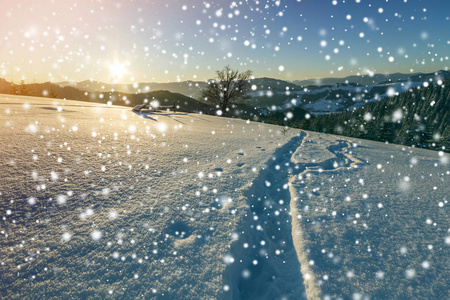 黎明时的冬季圣诞景观。 人类足迹轨迹路径在晶莹洁白的深雪中穿过空旷的田野，木本，黑暗的山脉，大片的雪花在晴朗的蓝天上复制空间背景