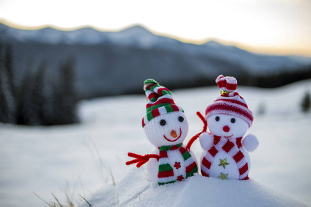 两个小有趣的玩具婴儿雪人在针织帽子和围巾在深雪户外模糊的山脉景观背景。 新年快乐，圣诞贺卡主题。