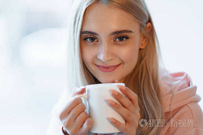 一个漂亮的微笑女孩坐在模糊的窗户玻璃旁，一边喝咖啡或牛奶。穿着一件随意的粉红色，苍白的帕卡。在靠近嘴唇的女人手中合上白色杯子。