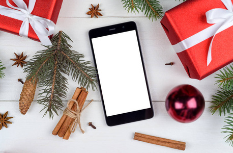 关闭移动智能手机与空白白色屏幕的应用程序演示与圣诞装饰复制空间。 圣诞礼物冷杉树枝在木制背景上。 平躺式顶部视图