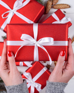 合上年轻女子的手，手里拿着白色木制背景上方的红色圣诞礼物。 带有红色礼品盒的圣诞背景。 平躺式顶部视图
