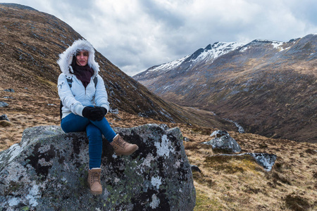 穿着白色蓬松夹克的女人坐在苏格兰内维斯山脉的岩石上，在一个多云的微风日。 白雪覆盖的山脉在后面。