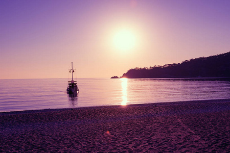 帆船在地中海的绿松石水域，在日落期间被岩石海岸包围，奥卢德尼兹是土耳其的旅游地标。 紫色的海景。 背光。