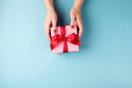 俯视女性手上拿着粉红色礼品盒，蓝色背景上用红色蝴蝶结包裹。 最小的风格成分。