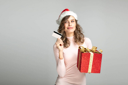 圣诞节购物的时间到了。 年轻漂亮的女人戴着圣诞帽，手里拿着信用卡和礼物，想着花钱