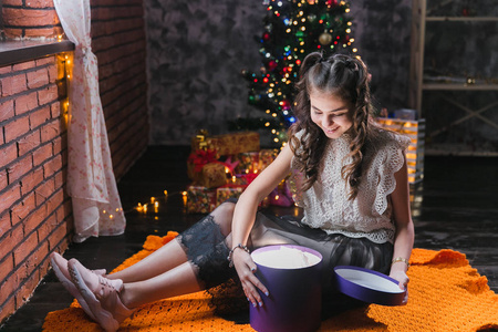 小女孩穿着连衣裙坐在圣诞树附近的地板上，拿着一个带礼物的盒子。小女孩坐在一个黑暗的房间里，打开一个带礼物的紫丁香盒子