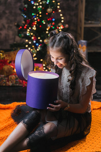 小女孩穿着连衣裙坐在圣诞树附近的地板上，拿着一个带礼物的盒子。小女孩坐在一个黑暗的房间里，打开一个带礼物的紫丁香盒子