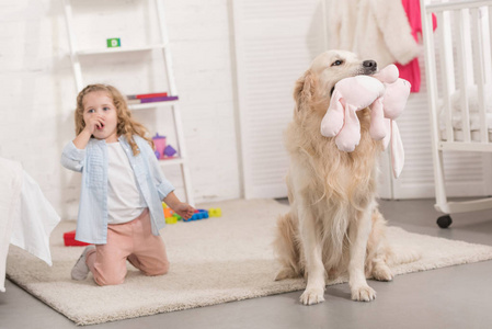 令人惊讶的是，可爱的孩子坐在金色猎犬旁边，拿着玩具在儿童房里