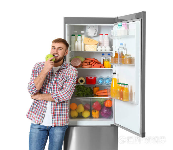 年轻人在白色背景的开放式冰箱旁吃苹果