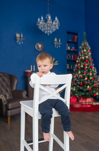 小男孩的肖像。 新年快乐。 装饰好的圣诞树。 圣诞节在明亮的蓝色房间。 坐在白色的椅子上