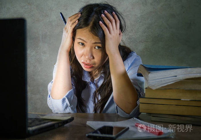 年轻的压力和沮丧的亚洲学生女孩努力工作,手提电脑和书堆在桌子上,不