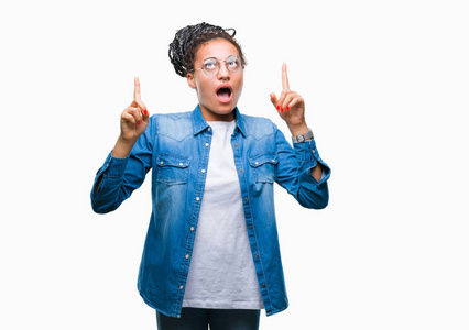 年轻的辫子非洲裔美国女孩戴着眼镜在孤立的背景下惊讶和惊讶地抬头，用手指和举起的手臂指着。