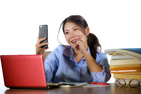 年轻美丽快乐的亚洲学生女孩，带着书堆和笔记本电脑桌，用手机自拍，微笑着，欢快地在白色背景下被隔离