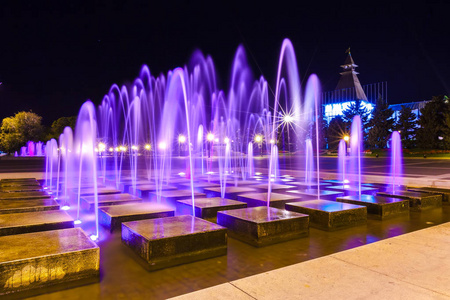 喷泉与紫色照明在列宁广场的中心与石头设计元素在走道内与绿树在黑暗的阿斯特拉罕克里姆林宫的背景。 晚上阿斯特拉罕城市俄罗斯。