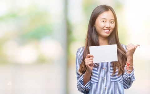 年轻的亚洲女人拿着空白卡片，在孤立的背景上指着，用拇指向旁边露出快乐的脸，微笑着