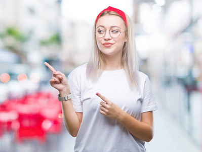 年轻的金发女人戴着眼镜，在孤立的背景上微笑，看着相机，双手和手指指向一边。