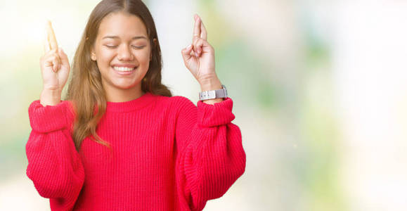 年轻美丽的黑发女人穿着红色的冬季毛衣，在孤立的背景上微笑着交叉手指，希望和眼睛紧闭。 运气和迷信的概念。