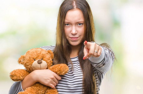 年轻的白种人女人拿着泰迪熊在孤立的背景上指着相机和你的手，从前面签署积极和自信的手势。