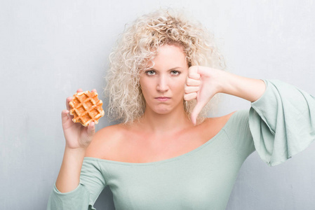 年轻的金发女人，在灰色的灰色背景下，吃比利时华夫饼，愤怒的脸，负面的迹象，显示不喜欢，拇指向下拒绝概念
