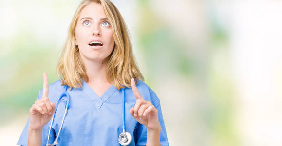 美丽的年轻医生女人穿着医用制服，在孤立的背景上惊讶和惊讶地抬头，用手指和举起的手臂指着。
