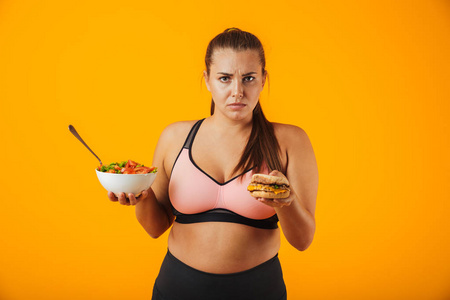 一位心情不好的超重健身女士的肖像，她穿着运动服，站在黄色背景下，端着一个碗，盛着沙拉和一个汉堡