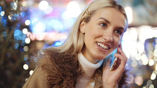 欧洲圣诞节市场上使用智能手机的女人。 圣诞节社交网络和交流的概念。 背景上模糊的圣诞灯。