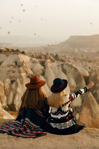 旅行。 女旅行者看着天空中飞舞的热气球，戴着帽子的女性坐在山上享受日落。 决议草案