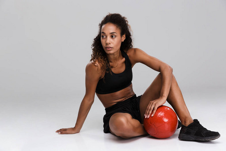 一位美国黑人年轻健美运动员的肖像，她用重球做运动，被隔离在灰色背景上，坐着看着远处