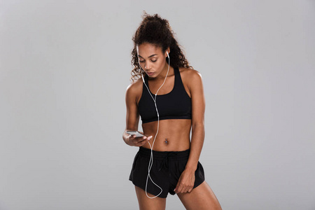 一位非洲裔美国年轻女运动员的肖像被隔离在灰色背景下，用耳机拿着手机听音乐