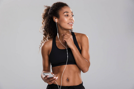 一位美国黑人快乐的年轻女运动员的肖像被隔离在灰色背景下，用耳机拿着手机听音乐