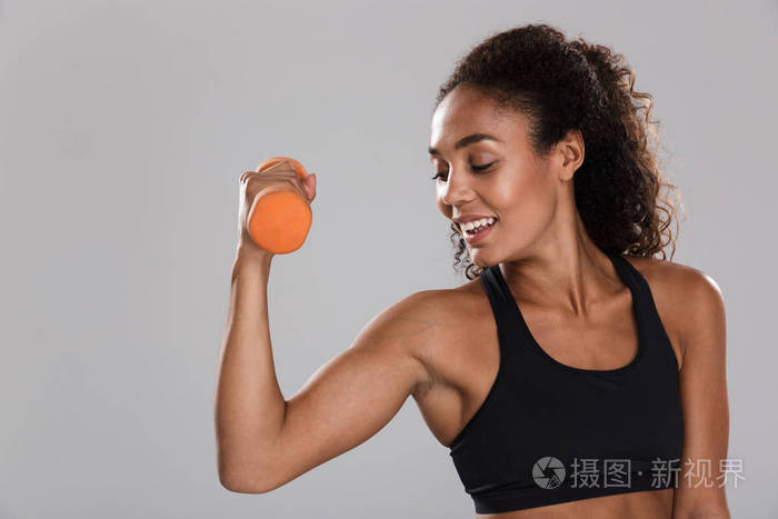 一位美国黑人微笑女运动员的肖像被隔离在灰色背景下，用哑铃锻炼