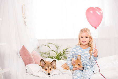 带着微笑的孩子坐在床上，家里养着彭布罗克威尔士科吉狗，手里拿着心形气球